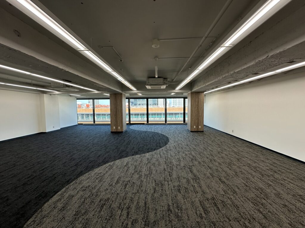 Empty, empty-office space with exposed ceiling pipes, long fluorescent light fixtures, and a row of large windows showing city buildings outside.
